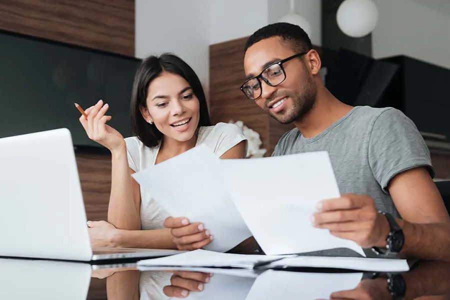 Annuities - Cheerful Young Couple Using a Laptop Together at the Kitchen Table While Analyzing Financial Documents