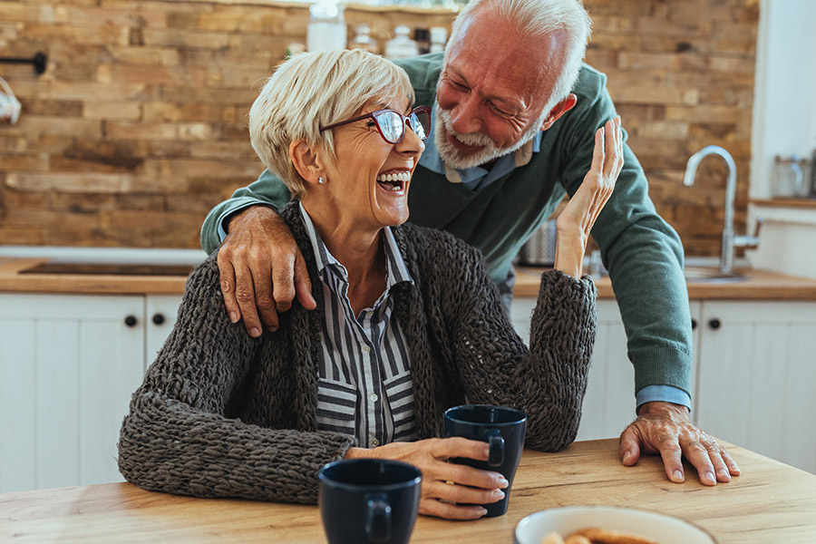 Medicare - Older Couple Smiling and Laughing in Their Kitchen While the Woman Sits at the Table and the Man has His Arm Around Her